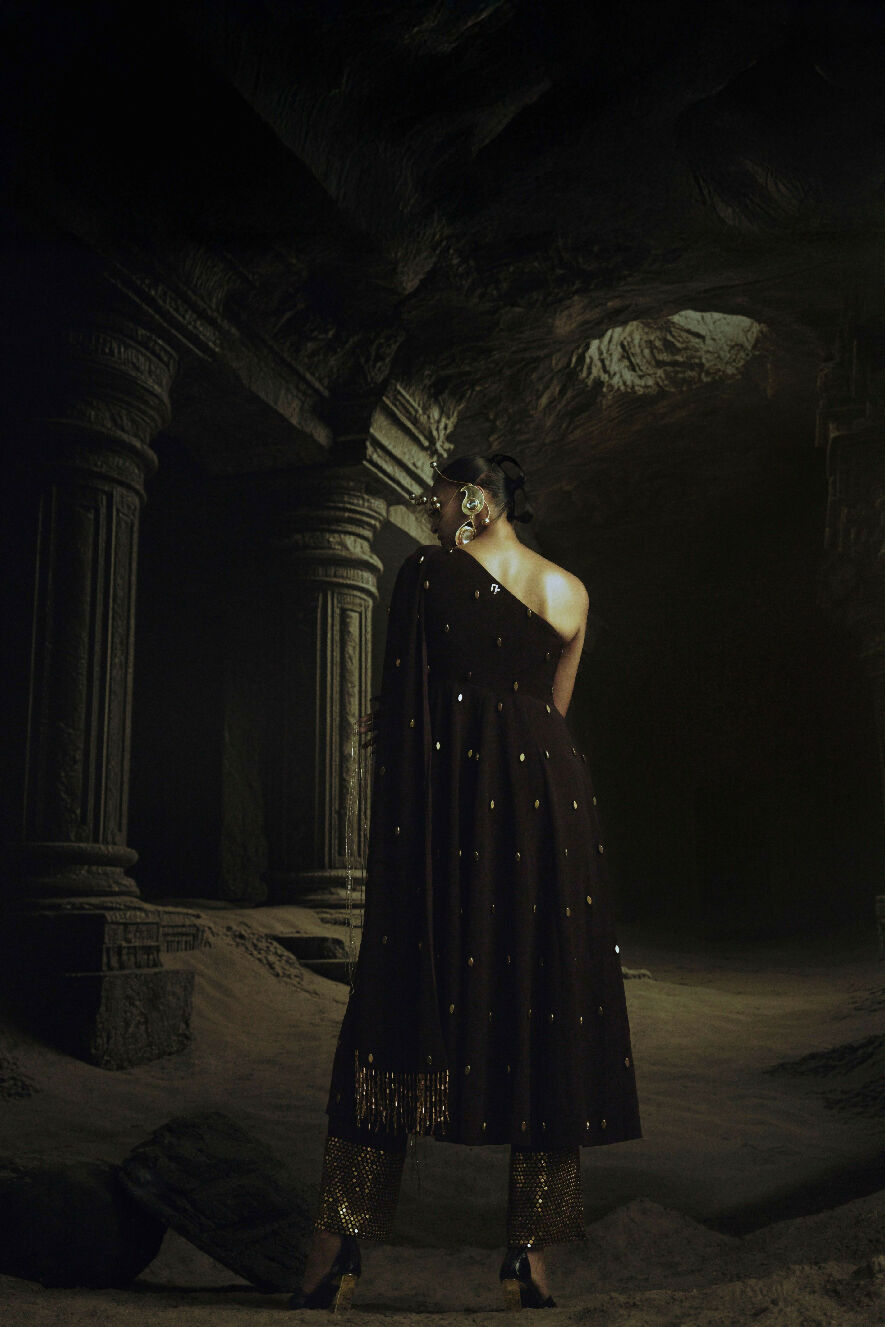 Person in a dark, ornate dress standing in a dimly lit, cave-like setting.