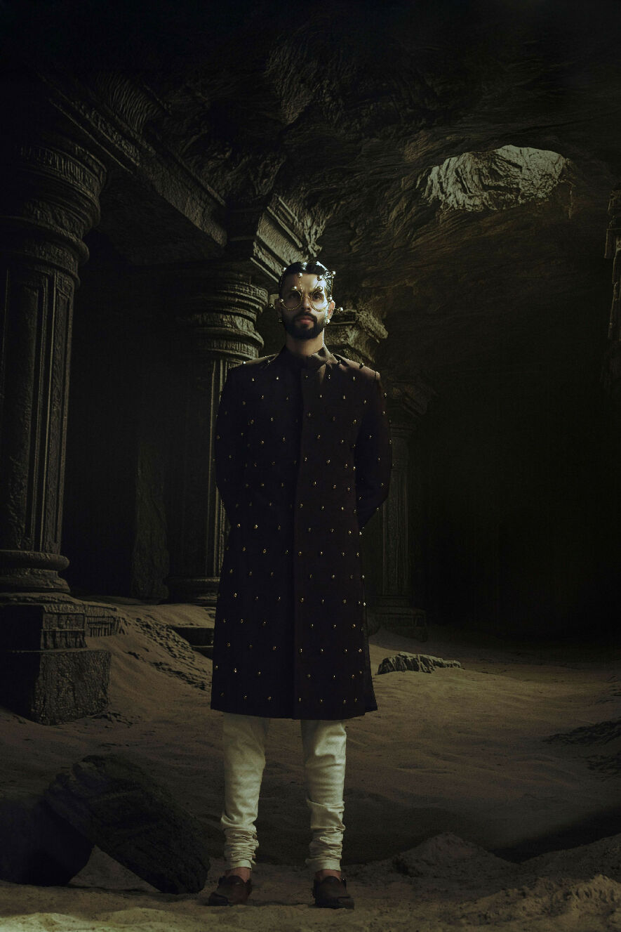 Man in a dark traditional outfit standing in a dark, possibly ancient setting with stone architecture.