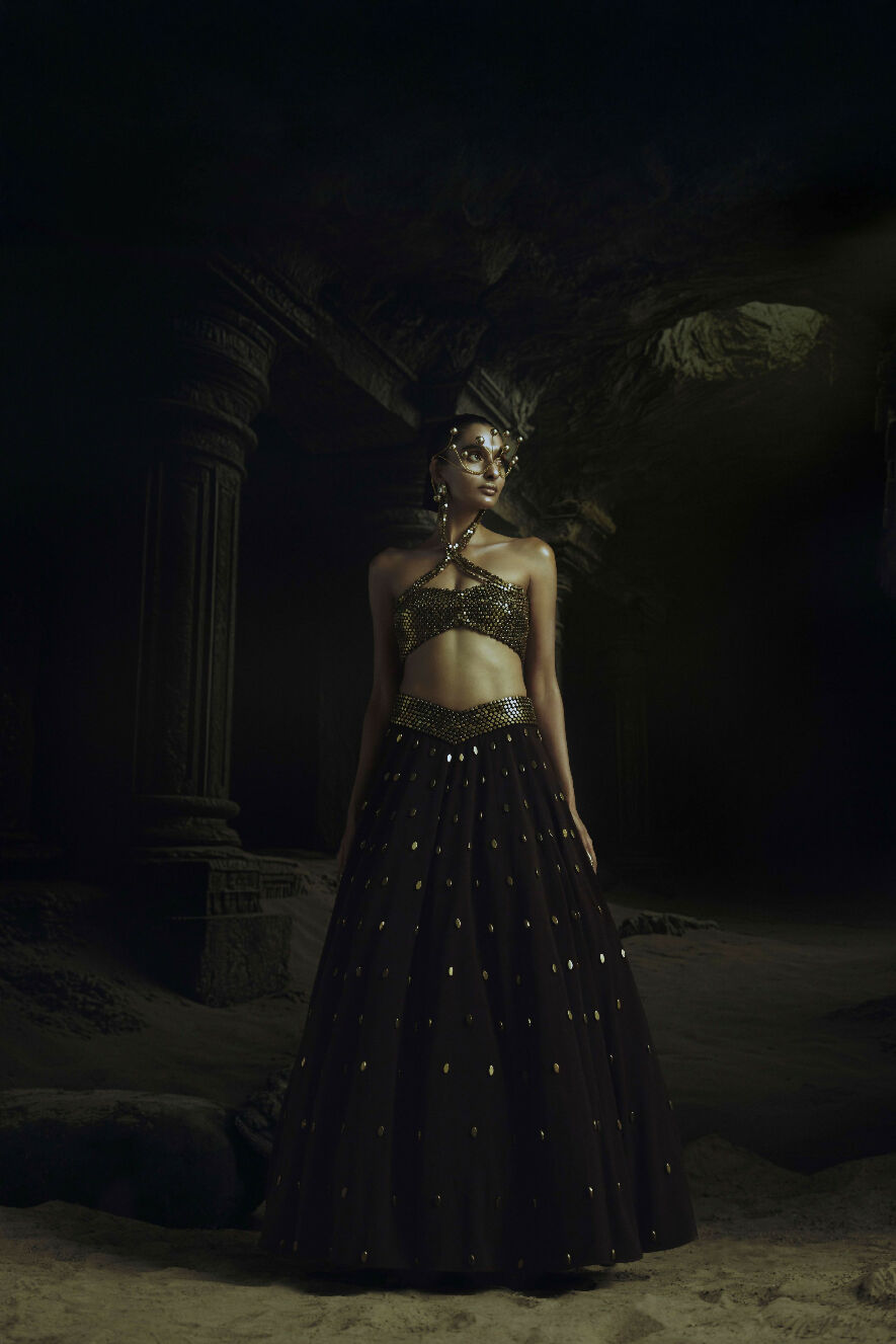 Woman in a dark, ornate dress standing in a dimly lit, cave-like setting.