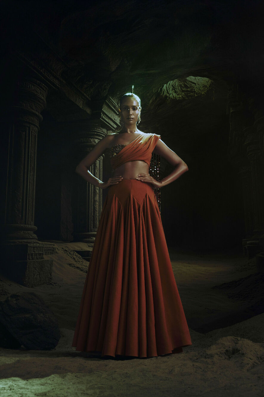 Woman in a red and orange outfit standing in a dark, cave-like setting.