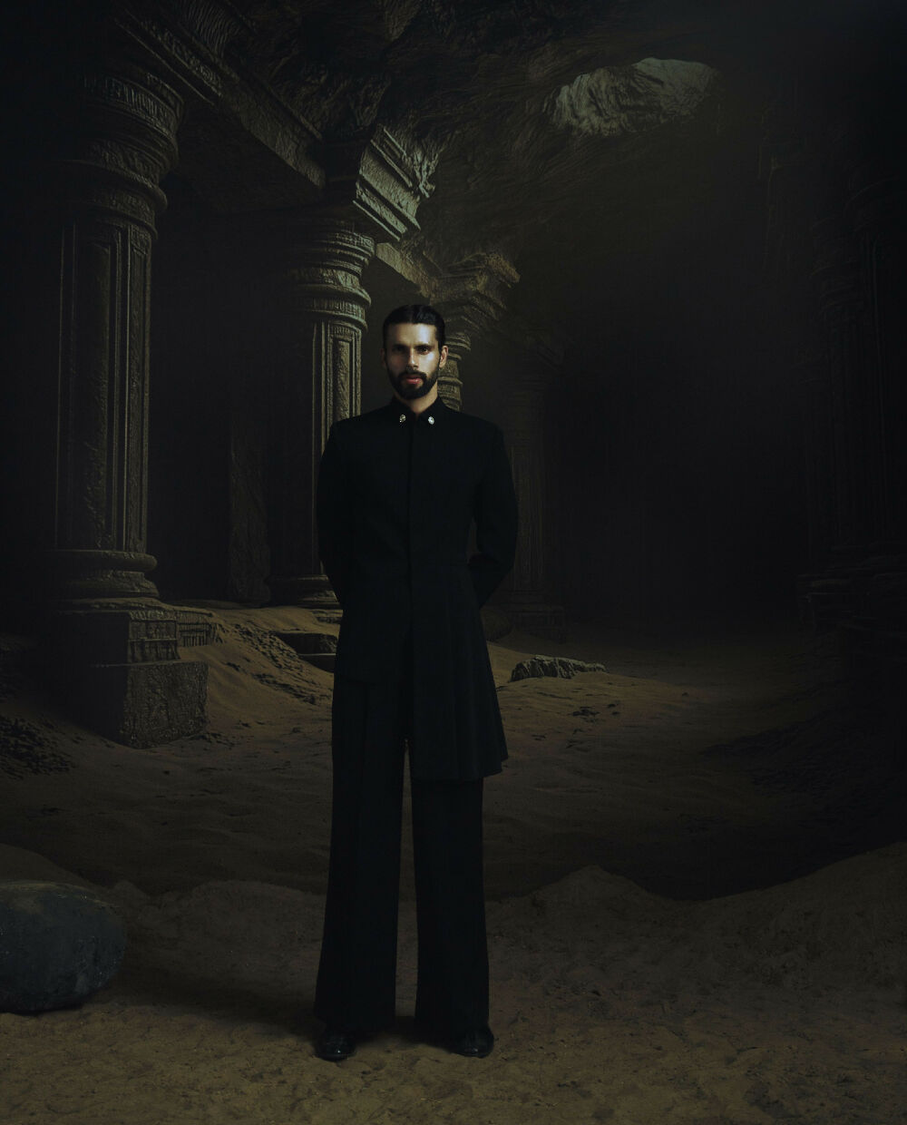 Man in a black suit standing in a dark, ancient ruin.
