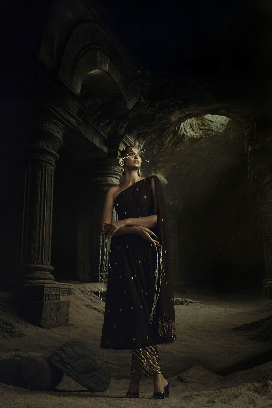 Woman in an elaborate dark dress with gold accents standing in a dimly lit, ancient ruin setting.