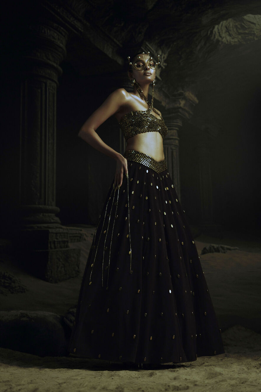 Woman in a dark, ornate Lehenga standing in a dimly lit, possibly ancient setting.