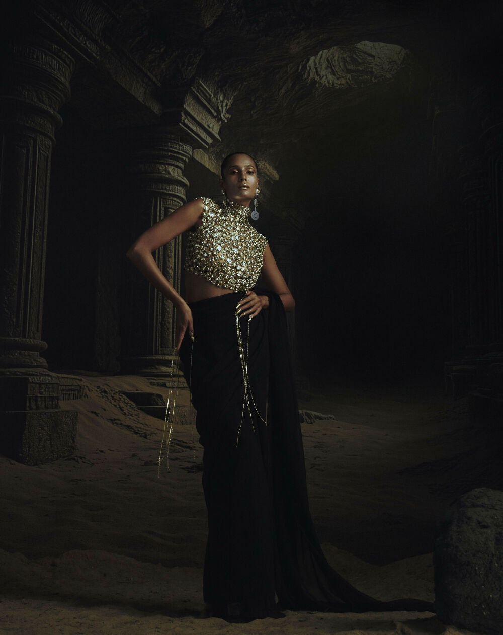 Woman in a sparkling top and black skirt standing in a dark, ancient temple setting.