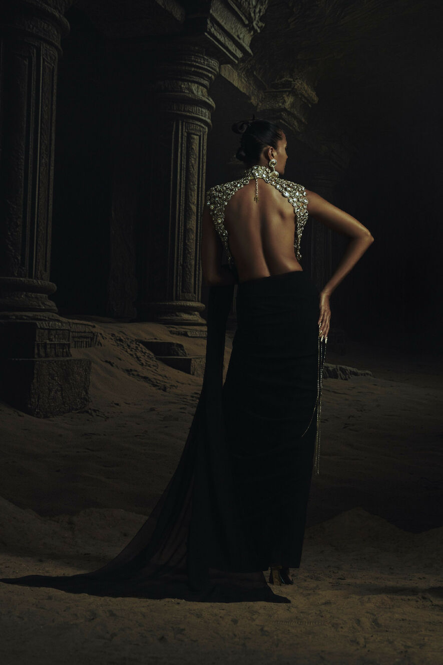 Woman in a black evening gown with a detailed back design in a dark, possibly ancient setting.