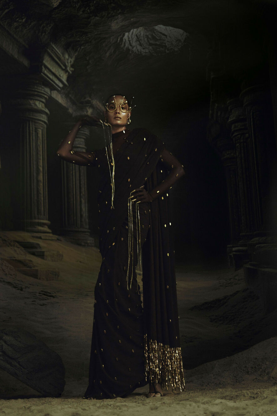 Woman in a black saree with gold details standing in a dark, ancient temple setting.