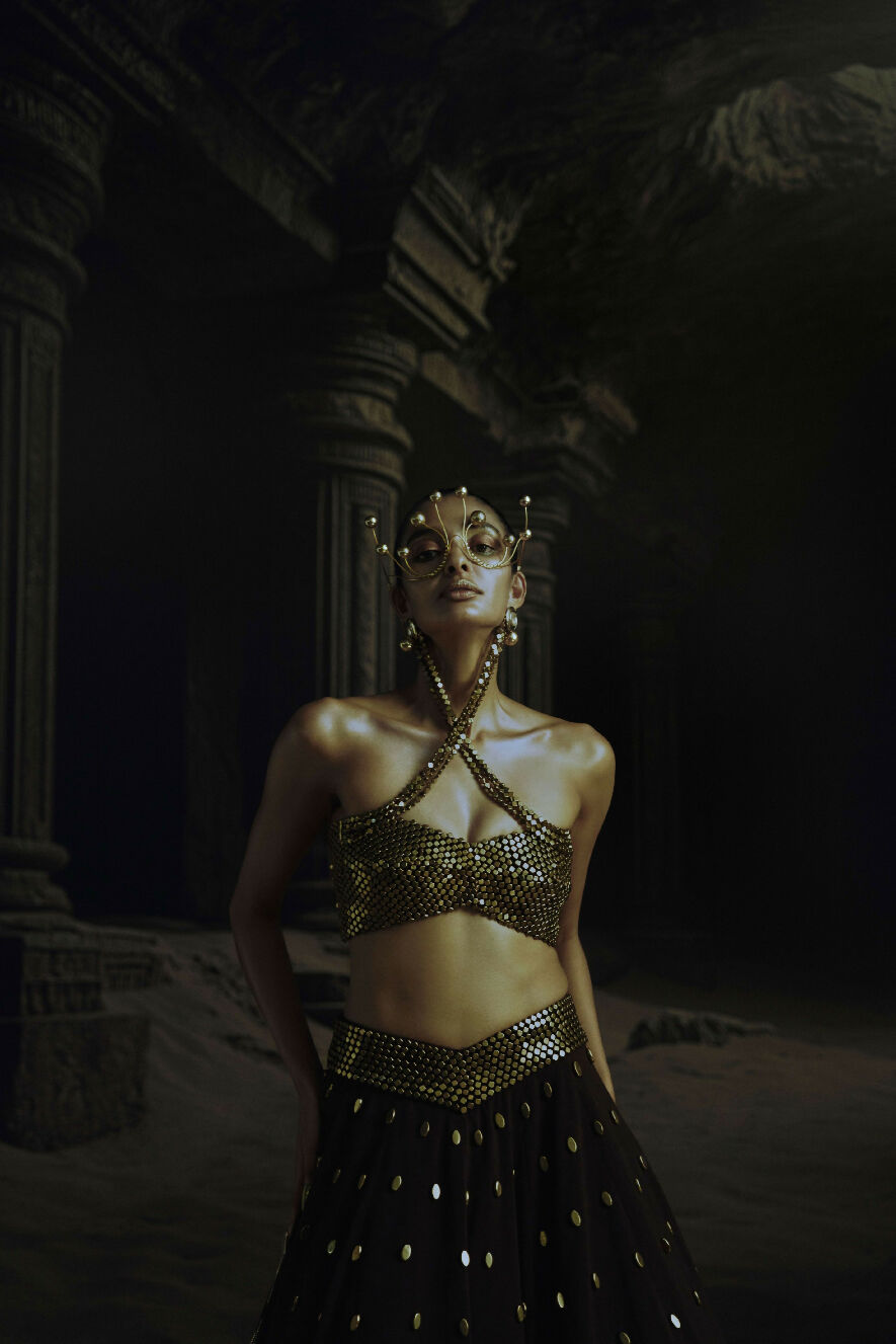 Woman in a gold and black Lehenga standing in a dark, desert-like setting with architectural columns.