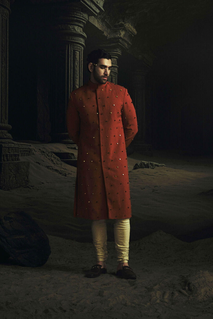 Man in a red traditional outfit standing in a dark, possibly ancient setting.