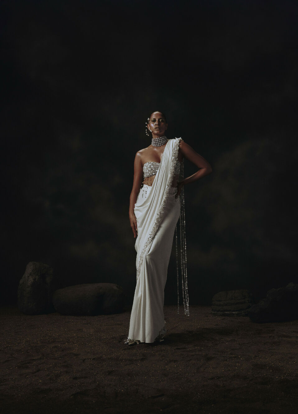 Woman in a white saree with intricate designs standing against a dark background