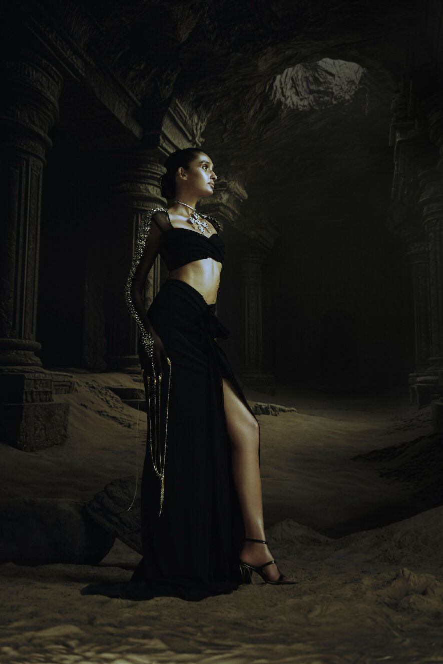 Woman in a dark, elaborate Kundan standing in a dimly lit, ancient ruin-like setting.